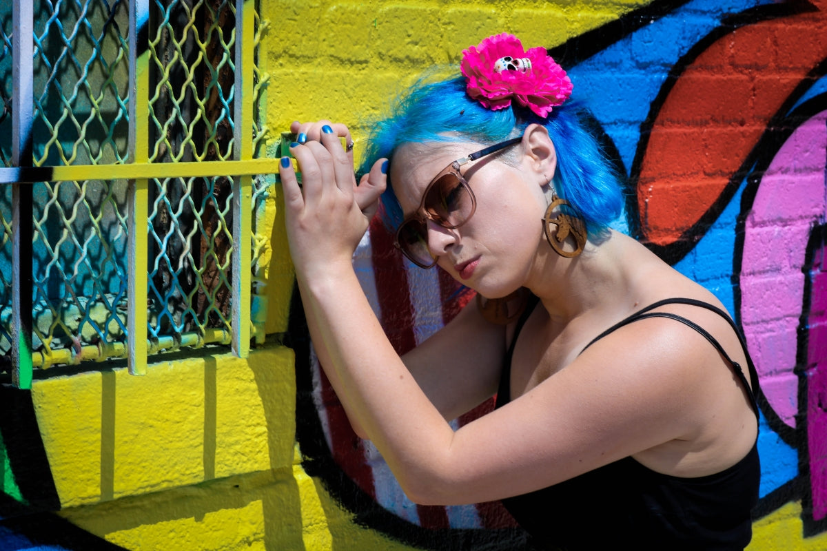 A non-binary individual with blue hair, pink hair clip, and sunglasses is standing in front of painted wall with their eyes closed